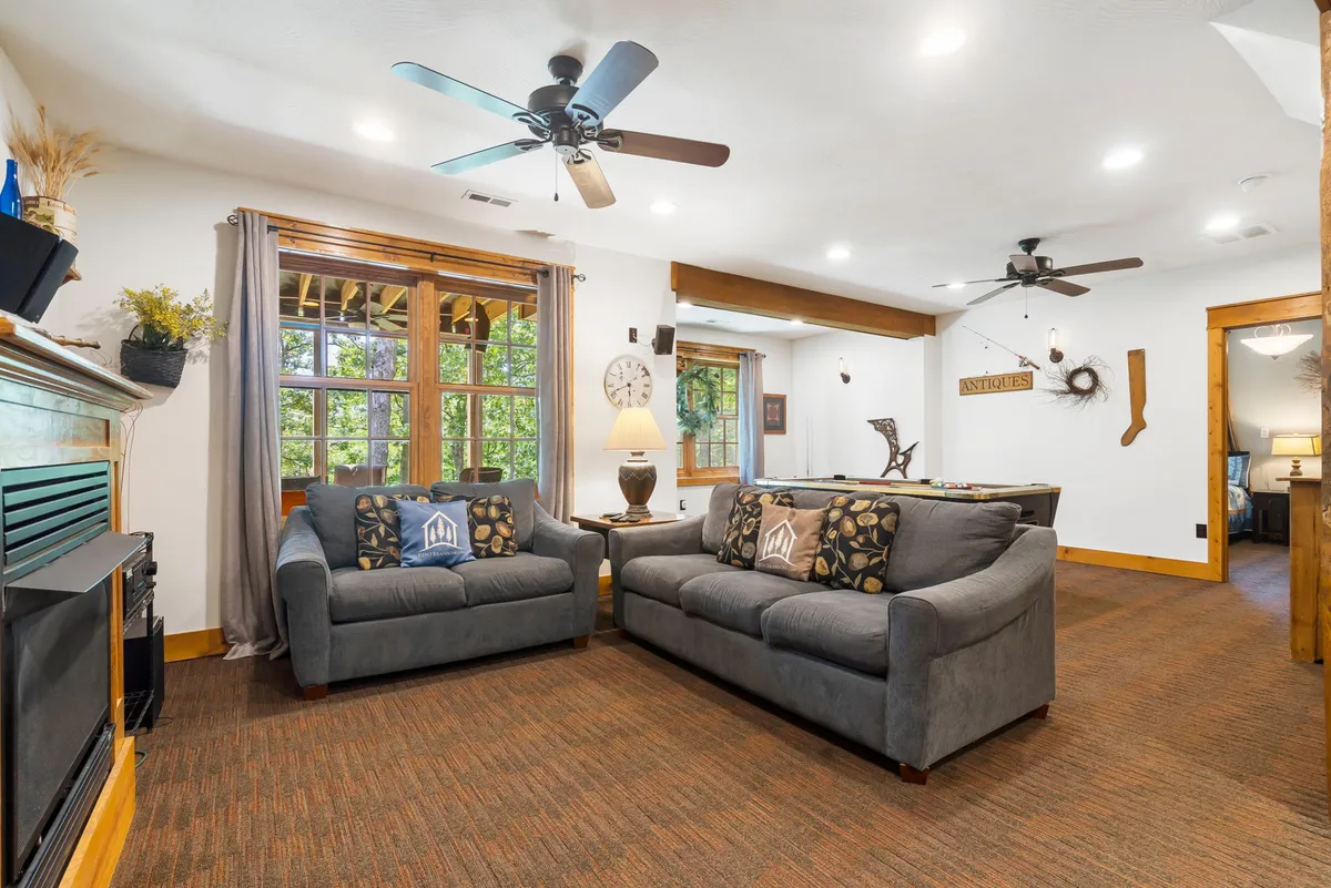 A cozy family room featuring comfortable sofas, decorative pillows, a prominent fireplace, large windows with views of the outdoors, and a table lamp providing ambient light. The arrangement is ideal for family game nights or relaxing together by the fire.