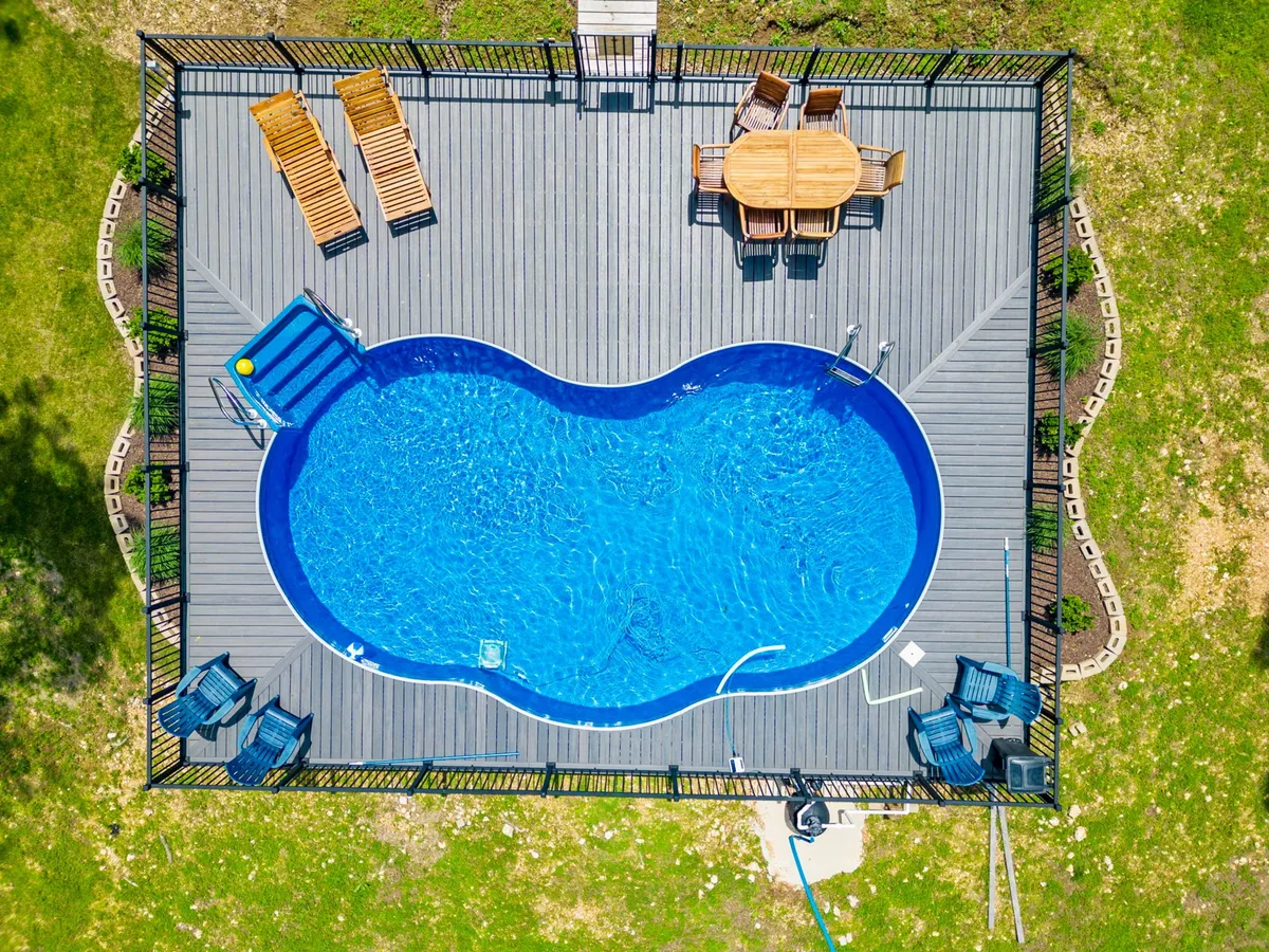 A sparkling swimming pool is surrounded by a spacious deck, furnished with lounge chairs and a shaded outdoor dining table. The area is bordered by lush greenery, creating a tranquil environment ideal for swimming, sunbathing, socializing, or hosting group activities.