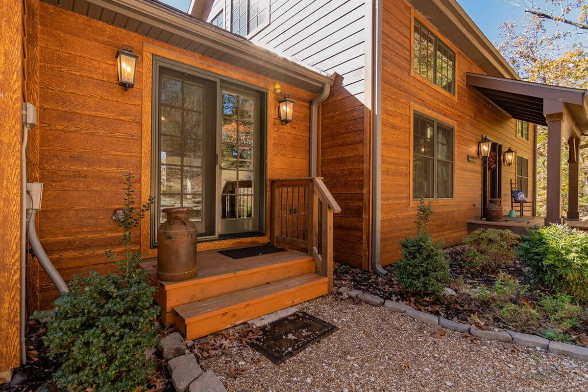 A welcoming exterior shot featuring a rustic wooden front door, staircase, and decorative lanterns framed by lush green shrubs. The porch suggests a comfortable and eco-friendly entrance, inviting guests to relax and enjoy the surrounding nature.