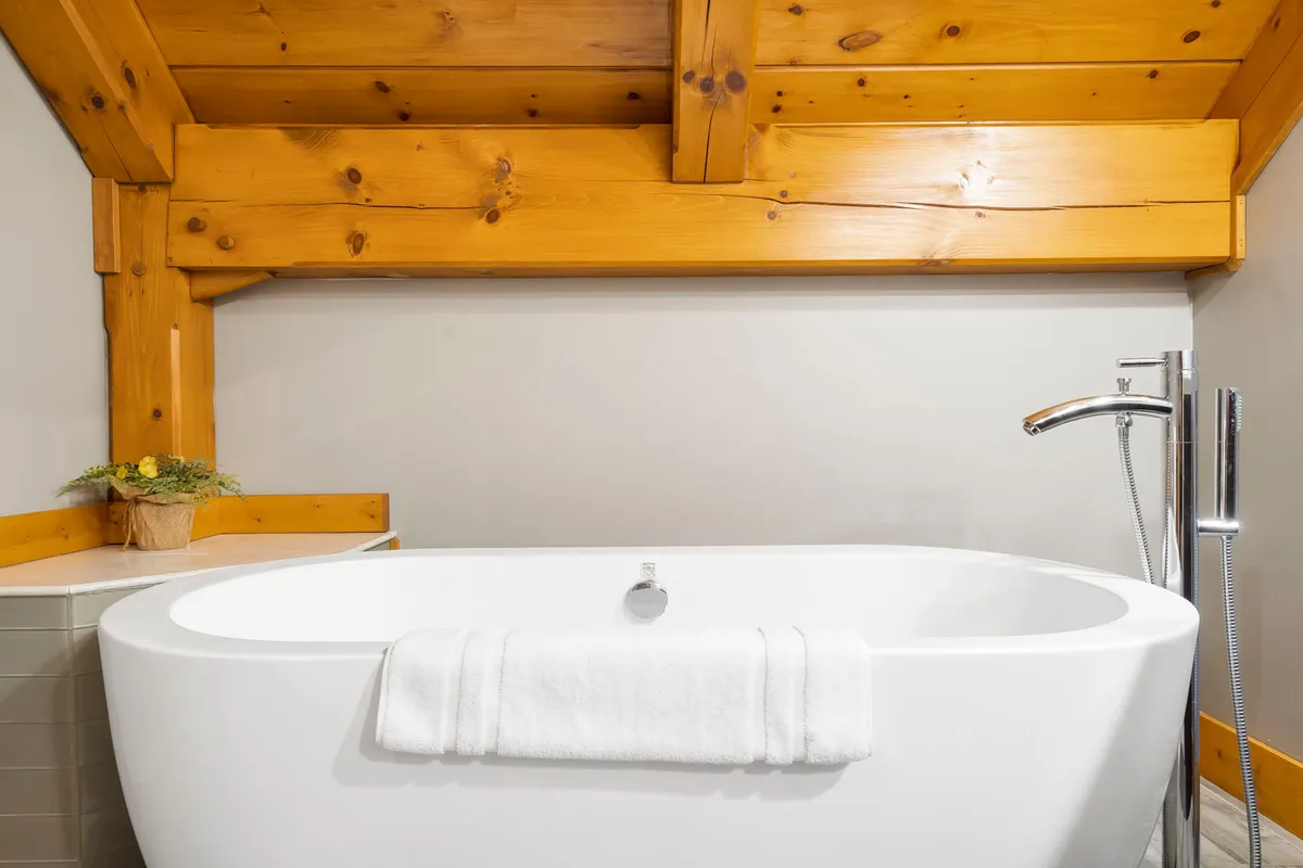 A luxurious bathroom with a freestanding soaker bathtub set against rustic wooden beams and accented with a decorative plant. The space is tranquil, bathed in soft natural light, and designed for relaxation and self-care.