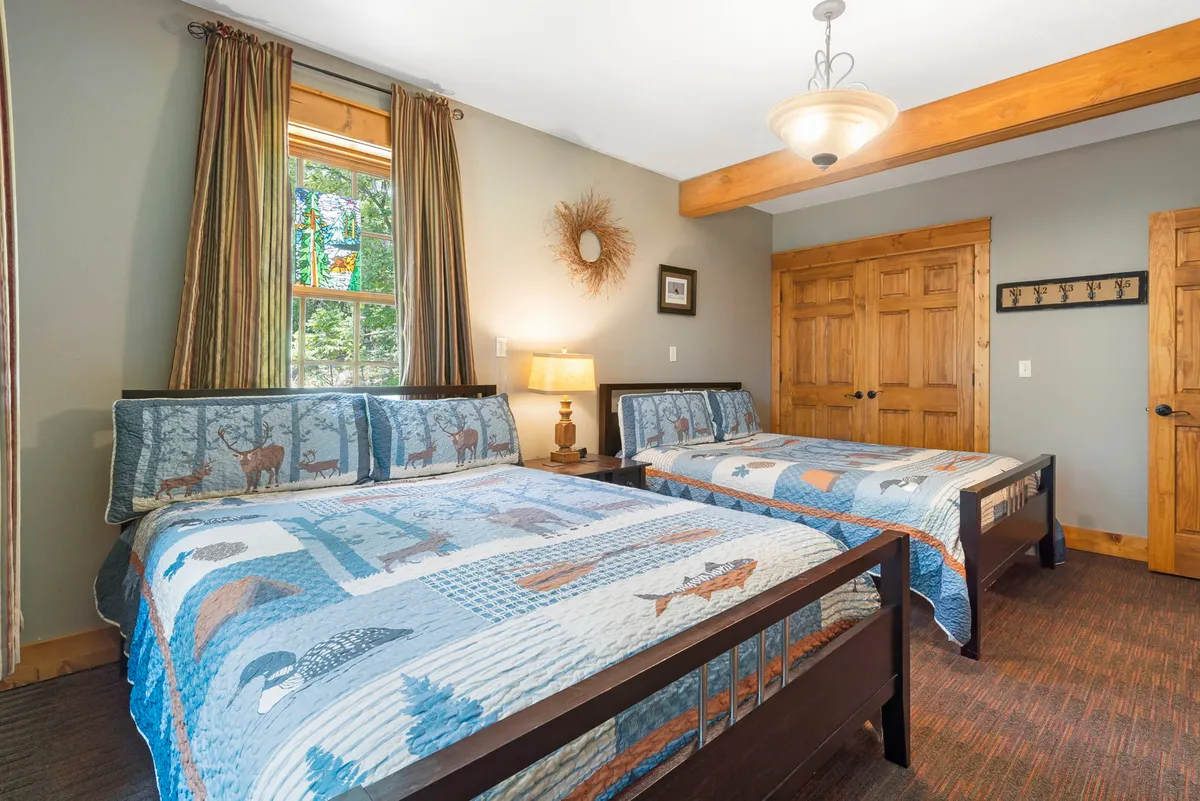 A cozy lower-level bedroom with two beds adorned in patterned quilts, a window allowing natural light to fill the space, wooden furniture, a lamp on the nightstand, and inviting wall art. The room feels welcoming, perfect for kids, teens, or friends.