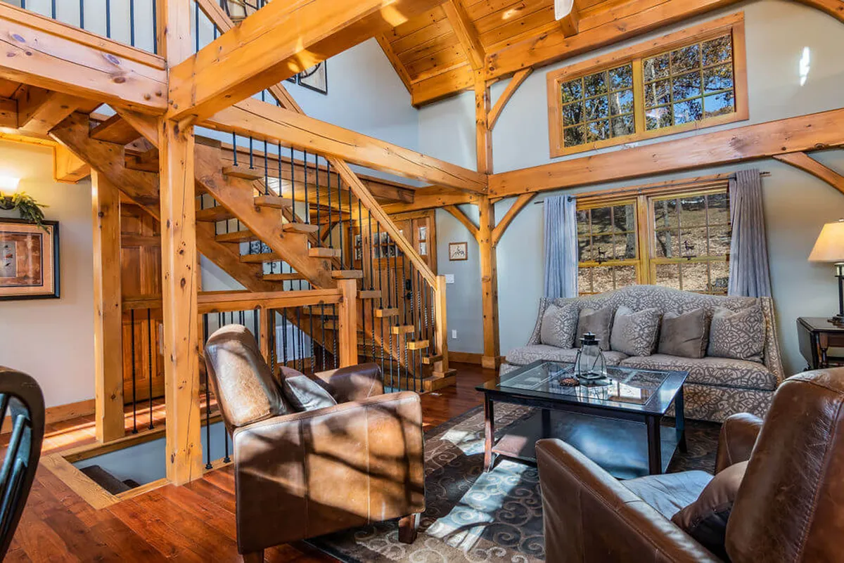 The image showcases a warm, inviting interior with a striking wooden staircase crafted from timber sourced on the property, large sun-filled windows revealing scenic woodland views, and a comfortable seating area with a rustic coffee table that invites gathering and relaxation. Soft natural light spills across the open space, highlighting the beautiful craftsmanship and cozy ambiance.