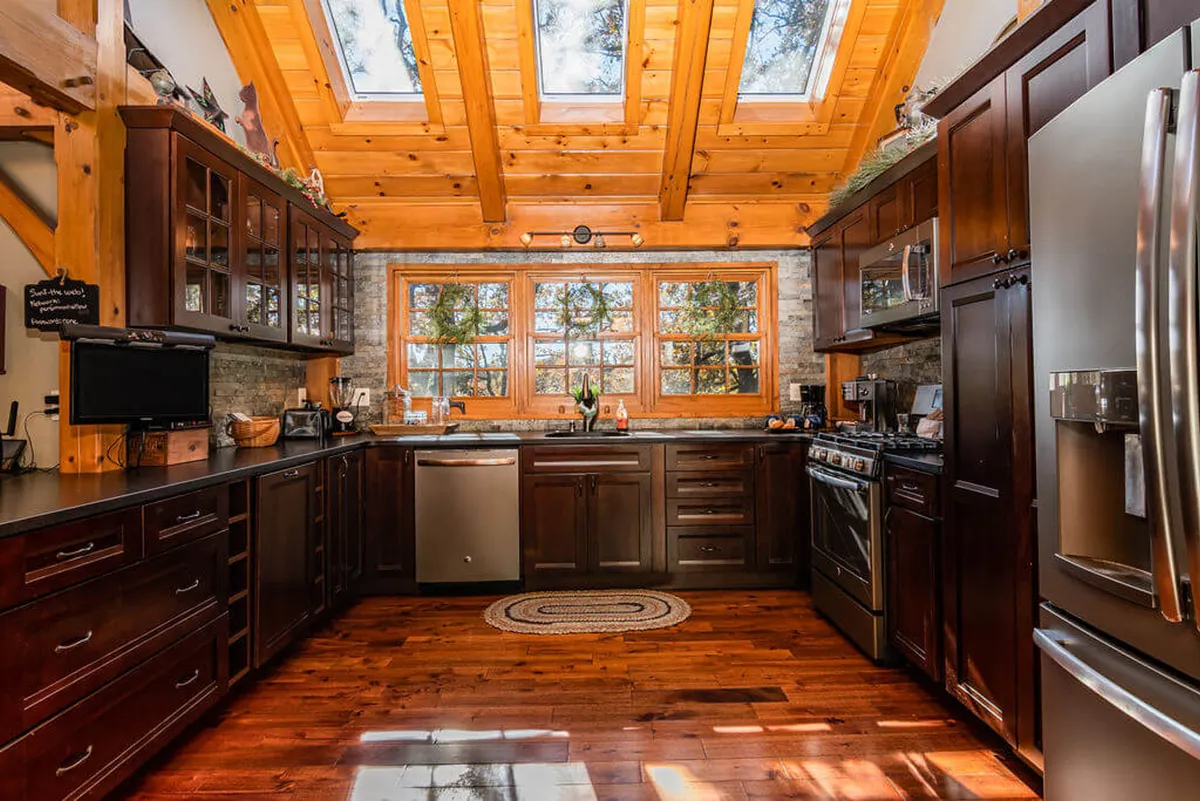 A beautiful kitchen scene with large windows, skylights, striking wooden ceiling beams, and dark cabinetry, where the kitchen serves as a bright, welcoming hub for cooking and socializing. The space looks ready for shared culinary experiences and family gatherings.