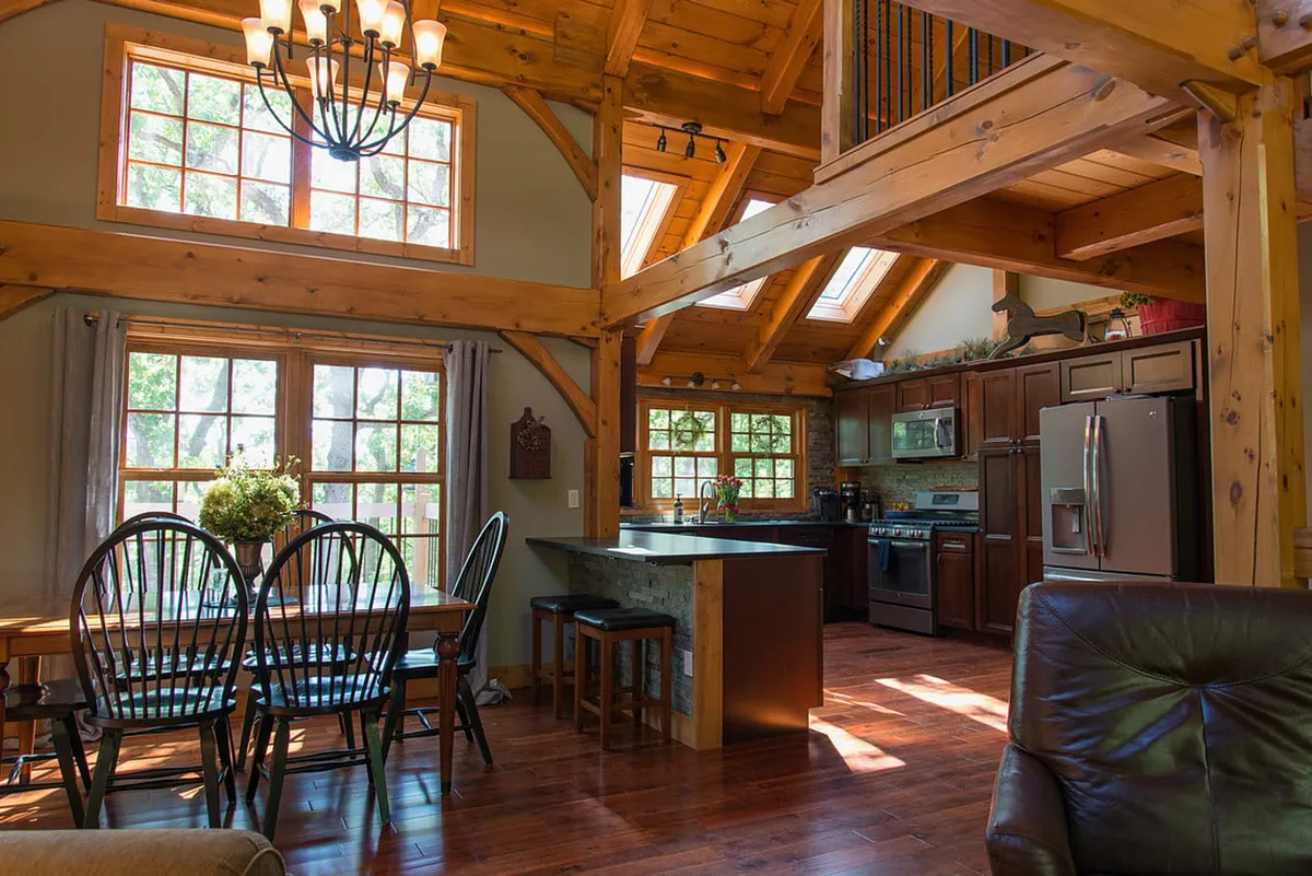 An open-concept kitchen and dining area with a large wooden dining table, kitchen island, exposed wooden beams, and ample natural light pouring through wide windows. The space is set for family meals and entertaining, with a seamless flow between cooking and dining areas.