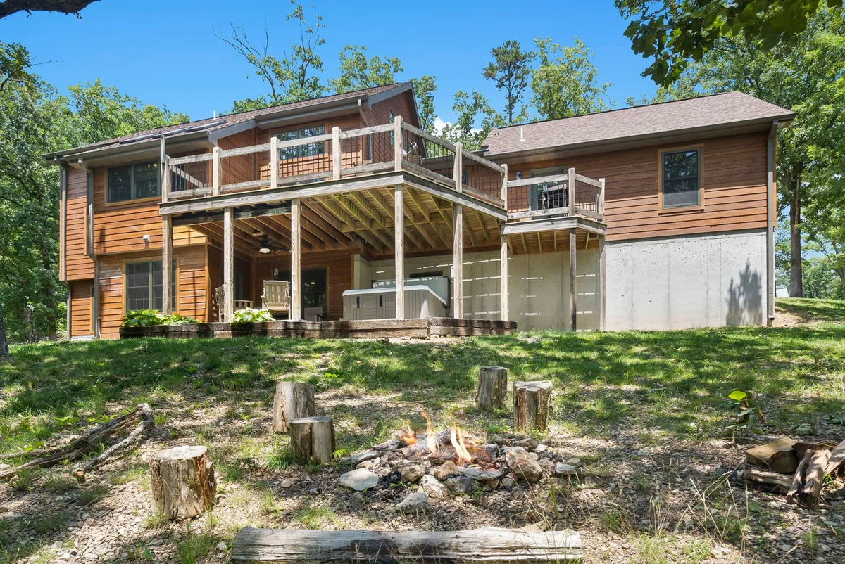 The image shows a deck with a fire pit, hot tub, and wooden structure, set among trees. It's an ideal spot for gathering around a fire, barbecuing, or relaxing in the hot tub while immersed in nature.