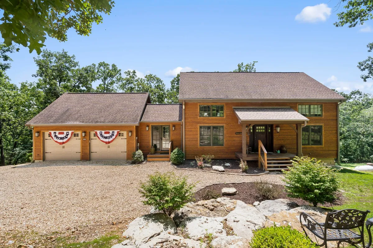 A full exterior view of the property’s wooden facade with a front porch, landscaped garden, and garage adorned with decorative banners. The scene offers a glimpse into outdoor family gatherings and a seamless connection to the natural surroundings.