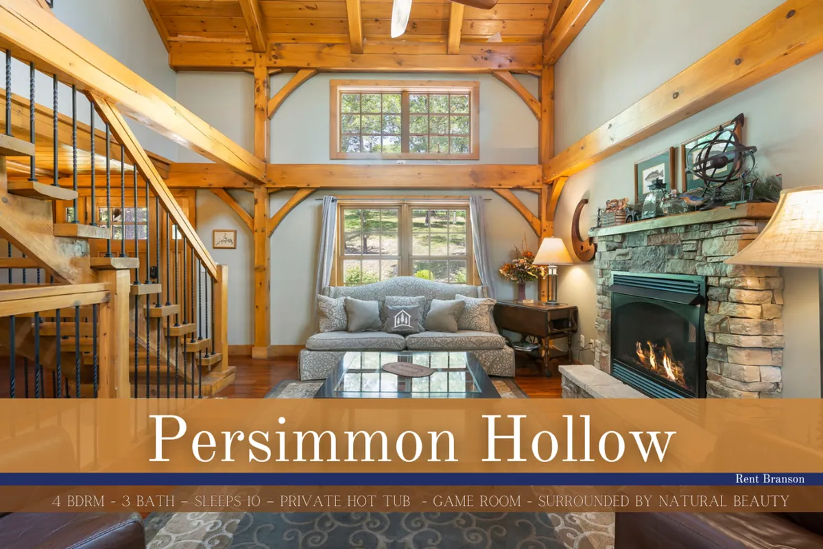 A spacious living room featuring a striking stone fireplace as the centerpiece, with soaring wooden beams overhead, comfortable seating arranged for conversation or relaxation, and expansive windows that capture the wooded scenery outside. The room is bathed in natural light, creating a warm and inviting space for gathering.
