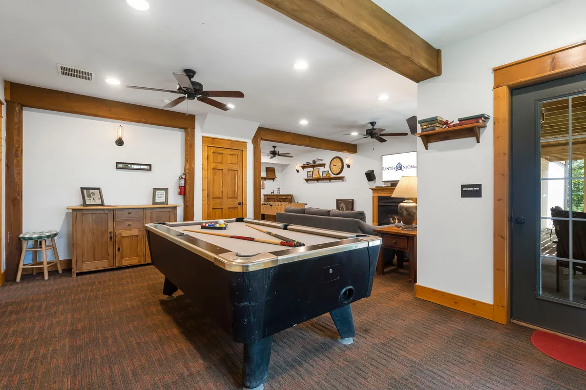 This image showcases a spacious game room featuring a pool table as the central focal point, surrounded by wooden cabinetry, a classic clock, a cozy seating area, and a warm fireplace. The relaxed, inviting atmosphere is perfect for socializing and recreational activities, while the visible TV hints at movie marathons and game nights.