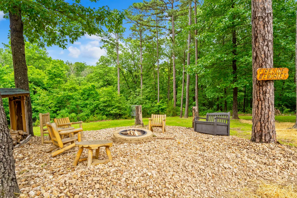 A cozy outdoor fire pit area surrounded by wooden chairs and a signpost, set amidst tall pines. The arrangement invites families to gather for evening storytelling, stargazing, and traditional campfire experiences.