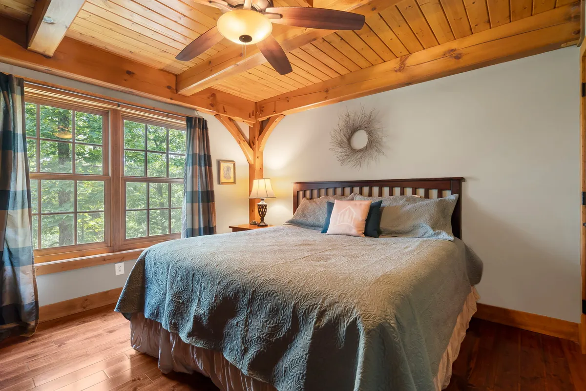 A tranquil bedroom featuring a king-size bed with crisp white linens, exposed wooden ceiling beams, and a large window offering a scenic view of the outdoors. The decor is simple yet elegant, exuding peace and comfort with an abundance of natural light streaming in.